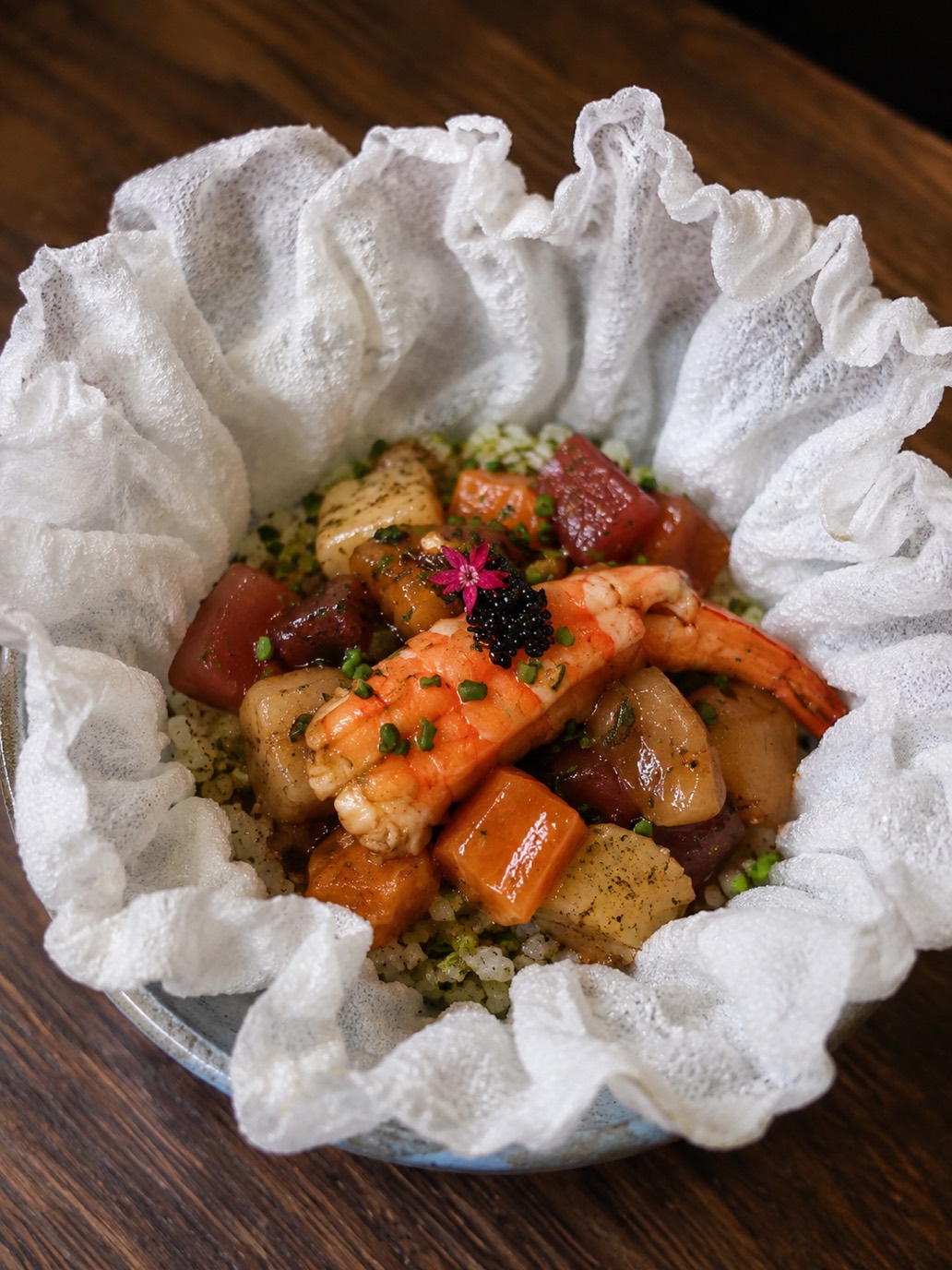 Tirashizushi servido em folha de arroz crocante em forma de flor: cubos de peixes frescos, camarão e ovas negras sobre shari com matcha, em bowl de cerâmica sobre madeira